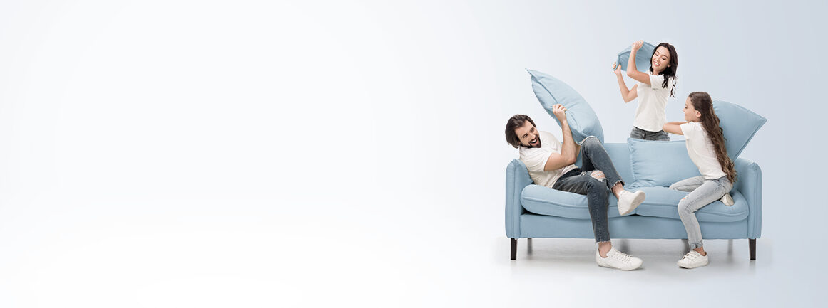 Eine dreiköpfige Familie liefert sich auf einem hellblauen Sofa eine spielerische Kissenschlacht. Der Vater hält ein Kissen abwehrend, während Mutter und Tochter, beide lächelnd, zum Angriff bereit sind. Sie befinden sich in einem minimalistischen, strahlend weißen Raum.