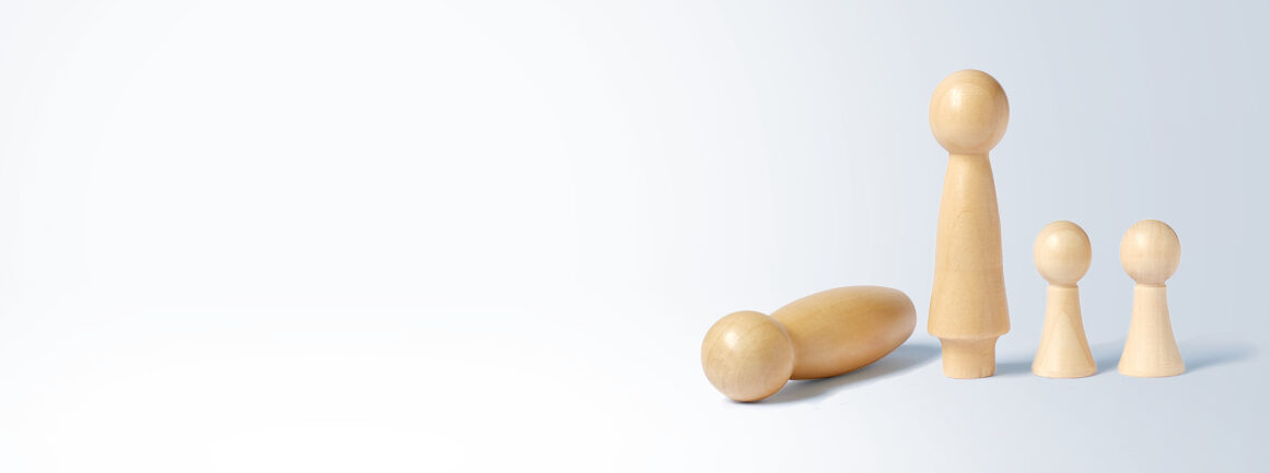 Vier Holzpuppen sind auf hellem Untergrund angeordnet. Eine große Puppe liegt auf der Seite, eine andere steht aufrecht. Zwei kleinere Puppen stehen daneben und bilden eine familienähnliche Gruppierung.