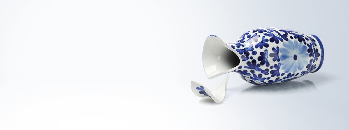 Eine blau-weiße Keramikvase mit Blumenmuster liegt auf der Seite, daneben liegt ein zerbrochenes Stück auf einer glatten, spiegelnden Oberfläche. Der Hintergrund ist ein weicher, heller Farbverlauf.