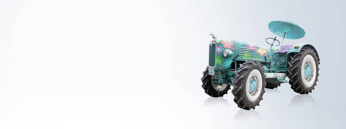 Ein Oldtimer-Traktor in leuchtenden Pastellfarben mit floralem Muster. Über dem Sitz ist ein kleiner, passender Sonnenschirm angebracht. Der Hintergrund ist leicht verfärbt und verleiht dem Bild eine verspielte Note.