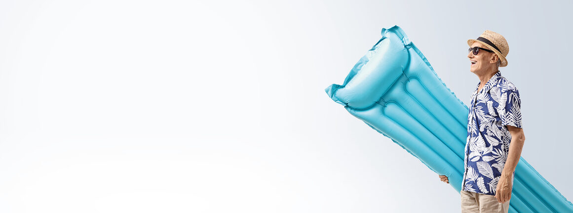Ein älterer Mann mit Strohhut und Tropenhemd hält vor einem hellen Hintergrund eine blaue aufblasbare Schwimmhilfe, lächelt und trägt eine Sonnenbrille.