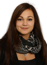 Eine Person mit langen, dunklen Haaren, einem schwarzen Oberteil und einem gemusterten Schal lächelt leicht in die Kamera. Der Hintergrund ist schlicht und unterstreicht das entspannte und zugängliche Auftreten der Person.