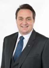 Ein Mann in dunklem Anzug, weißem Hemd und blauer Krawatte lächelt vor einem schlichten Hintergrund in die Kamera.