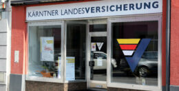 Das Schaufenster der Kärntner Landesversicherung besticht durch große Glasfenster mit einem farbenfrohen Logo. Der Eingang befindet sich mittig zwischen den Fenstern, in deren Inneren Plakate hängen. Die Außenfassade des Gebäudes ist rot-weiß gestrichen.