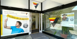 Eingang zu einer Filiale der Kärntner Landesversicherung. Auf dem Plakat eines Mannes in Blau steht „Flexibler als du denkst!“, rechts davon ein Cowboy-Display. Über der Tür prangt das Firmenlogo.