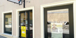Straßenansicht eines beigefarbenen Gebäudes mit großen Fenstern und einer Tür. An der Tür prangt eine gelbe Werbung, im Fenster hängen Plakate mit Versicherungsthemen. Darüber hängt ein schwarzes Metallschild mit Logo, das auf eine Versicherungsgesellschaft hinweist.