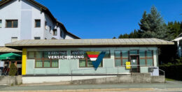 Ein Gebäude mit hellgrauem Dach und hellblauen Wänden trägt ein Schild mit der Aufschrift „Kärntner Landes Versicherung“ und einem bunten Logo. Es steht neben einer Straße unter einem strahlend blauen Himmel, in der Nähe ein Baum und ein weiteres Gebäude.
