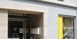 Das Bild zeigt den Eingang eines Versicherungsbüros mit einem Schild mit der Aufschrift „Kärntner Landes Versicherung“. Der Eingang verfügt über Glastüren, einige Briefkästen und ein gelb-blaues Fensterschild mit der gleichen Aufschrift.