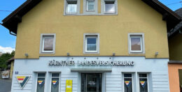 Ein gelbes Gebäude mit grauen Zierleisten trägt die Aufschrift „Kärntner Landesversicherung“. Die Fassade hat fünf Fenster und eine Tür. In den Fenstern sind dreieckige Logos mit blauen, gelben und roten Streifen zu sehen.
