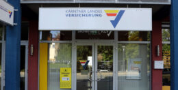 Schaufenster der Kärntner Landes Versicherung mit einem weißen Schild mit blauen und gelben Akzenten über Glastüren. Der Eingang ist mit verschiedenen Schildern versehen, und der Innenraum ist teilweise durch das Glas sichtbar.