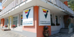 An einer Gebäudeecke ziert ein Schild mit der Aufschrift „Kärntner Landesversicherung“ ein V-förmiges Logo in den Farben Rot, Gelb und Blau. Das Gebäude hat große Fenster, eine rosa-weiße Fassade und einen Balkon mit weißem Geländer.