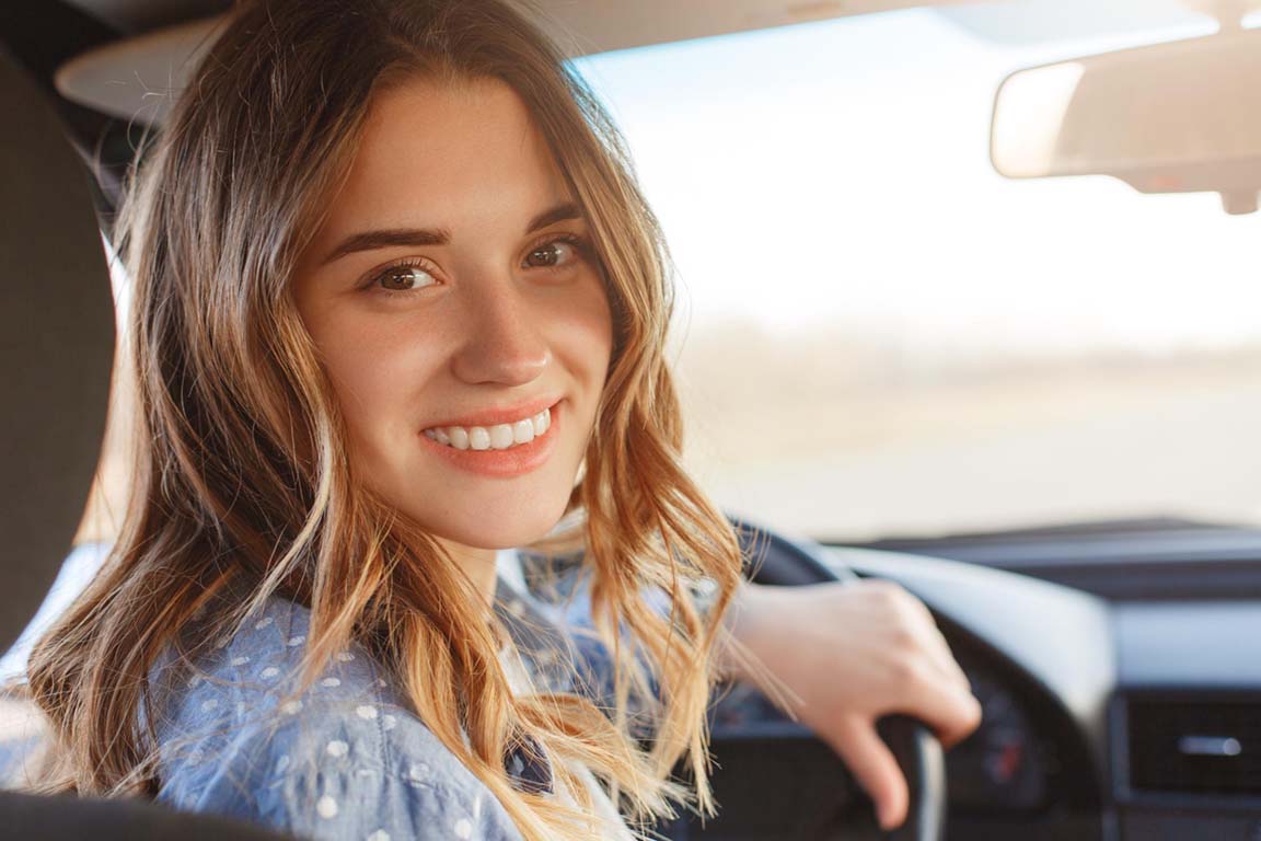 Eine Frau mit langen Haaren und einem blauen gepunkteten Hemd sitzt auf dem Fahrersitz eines Autos und lächelt in die Kamera. Der Innenraum des Autos ist sichtbar, und durch die Fenster fällt sanftes Tageslicht.