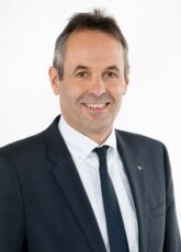 Eine Person in Anzug und Krawatte lächelt und steht vor einem schlichten weißen Hintergrund.