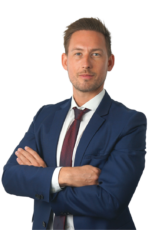 Ein Mann in blauem Anzug, weißem Hemd und roter Krawatte steht mit verschränkten Armen da und blickt direkt in die Kamera. Er hat kurze braune Haare und einen gepflegten Bart. Der Hintergrund ist schlicht weiß.