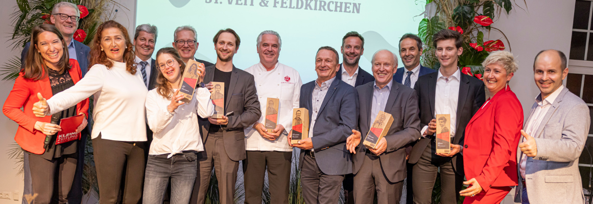 Eine Gruppe lächelnder Menschen posiert für ein Foto im Innenbereich. Einige halten hölzerne Auszeichnungen in den Händen. Sie sind in Business- und Freizeitkleidung gekleidet, stehen vor einem Hintergrund mit Text und sind von Pflanzen und Blumendekorationen umgeben.