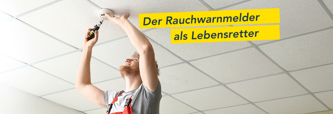Eine Person im Overall montiert mit einem Schraubenzieher einen Rauchmelder an einer weiß gefliesten Decke. Auf dem gelben Textschild steht „Der Rauchwarnmelder als Lebensretter“.