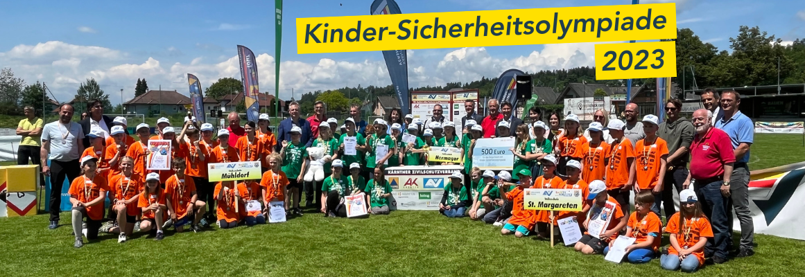 Eine große Gruppe von Kindern und Erwachsenen posiert auf einer Wiese. Die Kinder tragen bunte T-Shirts und halten Transparente und Urkunden in den Händen. Darüber steht auf einem Transparent „Kinder-Sicherheitsolympiade 2023“. Im Hintergrund sind Bäume und einige Gebäude zu sehen.
