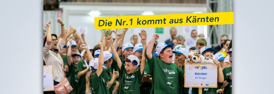 Eine Gruppe begeisterter Schulkinder in grünen Hemden und weißen Mützen jubelt und hält Schilder hoch. Über ihnen prangt ein Transparent mit der Aufschrift „Die Nr.1 kommt aus Kärnten“ – ein Hinweis darauf, dass sie aus Kärnten kommen. Die Stimmung ist festlich und ausgelassen.