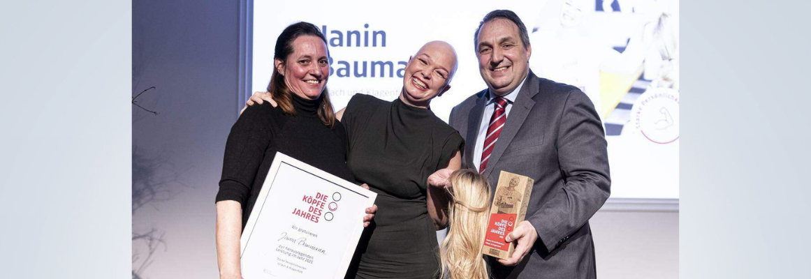 Drei lächelnde Personen bei einer Preisverleihung. Die Person in der Mitte hält eine goldene Auszeichnung, die Person links eine gerahmte Urkunde. Im Hintergrund wird auf einem Bildschirm Text angezeigt.