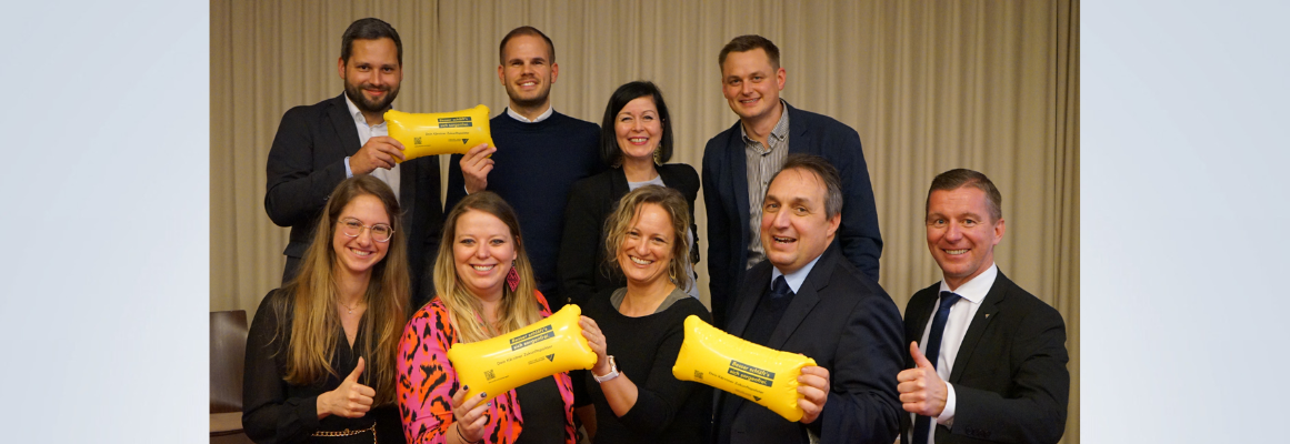 Eine Gruppe von acht Personen lächelt und hält gelbe Schecks in den Händen. Sie befinden sich in einem Raum mit einem beigen Vorhang im Hintergrund. Alle tragen Business- oder Freizeitkleidung, einige zeigen mit dem Daumen nach oben.