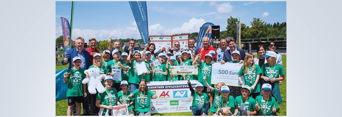 Eine Gruppe von Kindern und Erwachsenen posiert auf einem Sportplatz mit einem großen Scheck, Trophäen und Teddybären in den Händen. Die Kinder tragen grüne Hemden und Mützen. Im Hintergrund sind unter einem strahlend blauen Himmel verschiedene Banner und Flaggen zu sehen.
