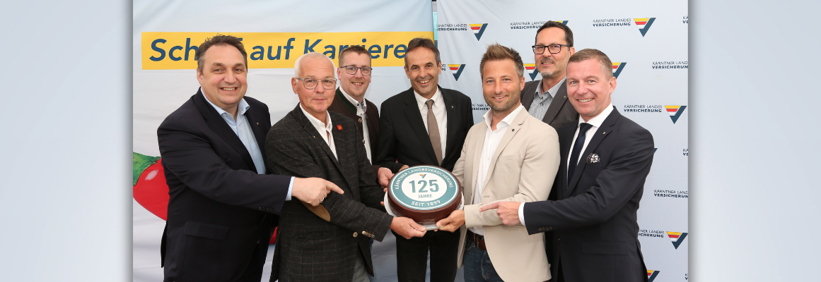 Sieben Männer in Anzügen stehen lächelnd nebeneinander und halten eine runde Tafel mit der Aufschrift „125 Jahre“ in den Händen. Vor ihnen stehen Banner mit Texten und Logos.