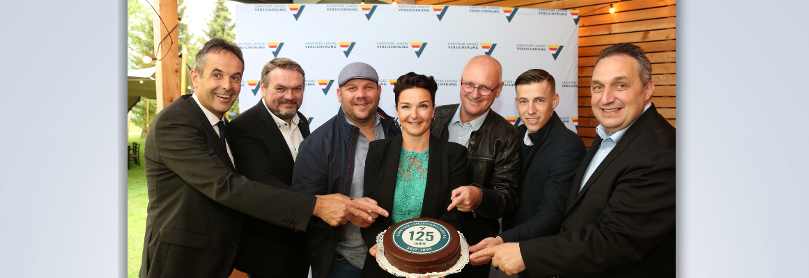 Sieben lächelnde Personen schneiden vor einem Hintergrund mit Logo und Text einen Kuchen mit der Aufschrift „125“ an. Sie tragen legere Business-Kleidung.