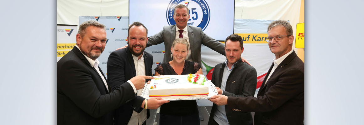 Sechs Personen stehen zusammen und halten eine große Torte mit einem Logo darauf. Sie lächeln und scheinen ein Ereignis zu feiern. Im Hintergrund sind ein Monitor und Banner mit Text zu sehen.