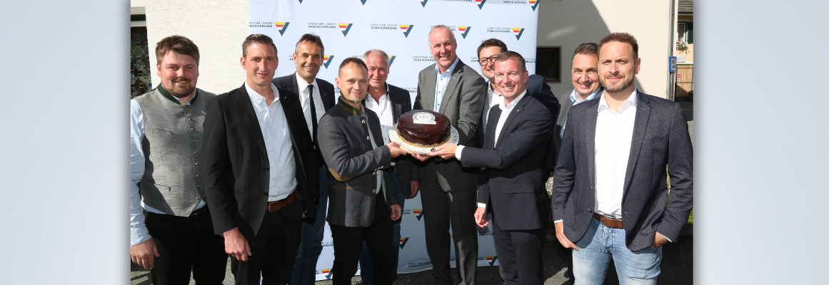 Eine Gruppe von zehn Männern in formeller Kleidung steht lächelnd vor einem Hintergrund mit Logos. Zwei von ihnen halten eine Torte mit der Zahl „175“. Die Gruppe scheint einen Anlass im Freien zu feiern, wobei das Sonnenlicht Schatten wirft.