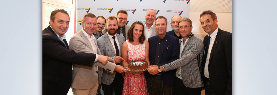 Eine Gruppe von Menschen steht im Halbkreis in einem Raum, lächelt und zeigt auf einen Schokoladenkuchen mit der Aufschrift „125“. Eine Frau in der Mitte hält den Kuchen. Im Hintergrund sind ein Logo und ein Design zu sehen.