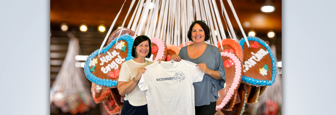 Zwei Frauen lächeln und halten ein weißes Shirt mit der Aufschrift „WIESNMARKT“ in den Händen. Sie stehen vor großen, herzförmigen Lebkuchen, die an den Wänden hängen.