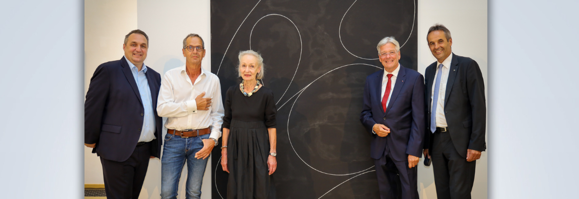 Fünf Personen stehen gemeinsam vor einem großen schwarzen Gemälde mit abstrakten weißen Linien. Sie tragen formelle und halbformelle Kleidung und lächeln in die Kamera. Die Szenerie erinnert an eine Kunstgalerie oder einen Ausstellungsraum.