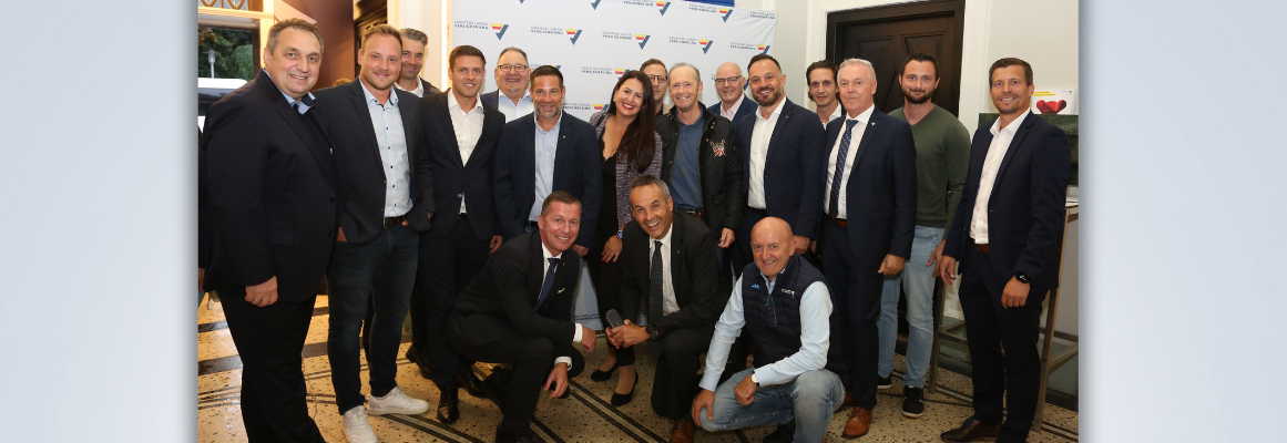 Eine Gruppe von Menschen in Business-Kleidung steht lächelnd für ein Foto zusammen. Sie befinden sich in einem Gebäude, hinter dem sich ein Markenhintergrund und darunter ein gemusterter Boden befindet. Einige knien vorn, andere stehen dahinter.