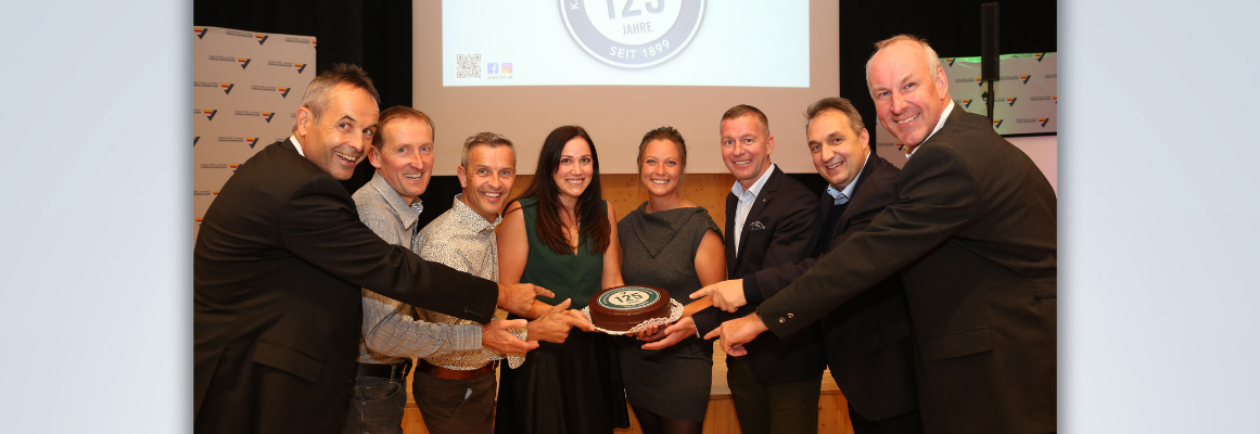 Eine Gruppe von acht Personen steht lächelnd im Halbkreis um eine Torte. Auf der Torte steht „125 Jahre“. Ein projiziertes Bild hinter ihnen zeigt denselben Text zusammen mit „seit 1898“.