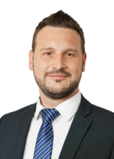 Ein Mann mit kurzen dunklen Haaren und Bart, der einen Anzug, ein weißes Hemd und eine blau gestreifte Krawatte trägt, blickt mit einem leichten Lächeln vor einem schlichten Hintergrund nach vorne.
