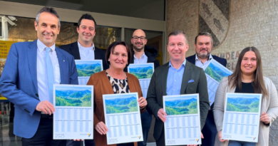 Eine Gruppe von sieben Personen in Business-Kleidung steht vor einem Gebäude und hält Kalender für 2024 mit malerischen Landschaftsmotiven in den Händen. Sie lächeln und stehen in zwei Reihen vor einer Fassade aus Glas und Ziegeln.