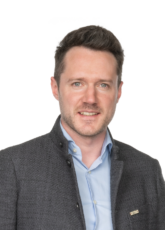 Ein Mann mit kurzen braunen Haaren, der ein blaues Hemd und einen dunklen strukturierten Blazer trägt, steht vor einem schlichten Hintergrund und lächelt sanft.