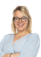 Eine Person mit blonden Haaren und Brille lächelt. Sie trägt eine hellblaue Langarmbluse und hat die Arme vor der Brust verschränkt. Der Hintergrund ist schlicht weiß.