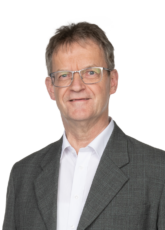 Ein Mann mittleren Alters mit Brille und kurzen Haaren, der ein weißes Hemd und ein dunkles Jackett trägt, lächelt vor einem schlichten Hintergrund in die Kamera.