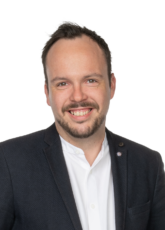 Lächelnder Mann mit kurzen Haaren und Bart, der einen dunklen Blazer über einem weißen Hemd trägt. Er hat einen freundlichen Gesichtsausdruck und steht vor einem neutralen Hintergrund.