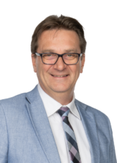 Lächelnder Mann mit Brille, hellblauem Jackett, weißem Hemd und gemusterter Krawatte vor schlichtem Hintergrund.