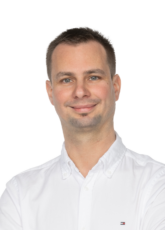 Ein Mann mit kurzen Haaren lächelt und trägt ein weißes Hemd mit Kragen. Der Hintergrund ist schlicht.
