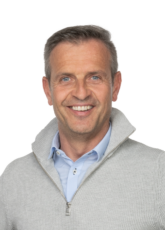 Lächelnder Mann in einem grauen Pullover mit Reißverschluss über einem blauen Hemd mit Kragen vor einem schlichten weißen Hintergrund.