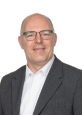 Ein Mann mit Brille, dunkelgrauem Sakko und weißem Hemd lächelt in die Kamera. Der Hintergrund ist schlicht weiß.