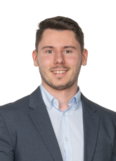 Ein Mann mit kurzen braunen Haaren und Bart lächelt in die Kamera. Er trägt einen blauen Blazer über einem hellblauen Hemd. Der Hintergrund ist schlicht weiß.
