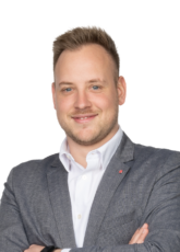 Lächelnder Mann in grauem Jackett und weißem Hemd, mit kurzen hellbraunen Haaren, steht mit verschränkten Armen da. Weißer Hintergrund.
