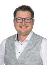 Lächelnder Mann mit Brille, weißem Hemd und gemusterter grauer Weste, posiert vor weißem Hintergrund.