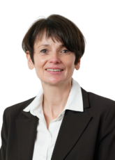 Eine Person mit kurzen braunen Haaren lächelt, trägt ein weißes Hemd mit Kragen und einen dunklen Blazer. Sie blickt nach vorne vor einem schlichten Hintergrund.