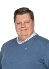 Eine Person, die einen blauen Pullover über einem weißen Hemd mit Kragen trägt, lächelt vor einem schlichten weißen Hintergrund in die Kamera.