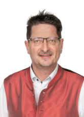 Ein Mann mit Brille und kurzen Haaren trägt ein weißes Hemd und eine rote, ärmellose Jacke. Er lächelt leicht und blickt direkt in die Kamera. Der Hintergrund ist schlicht weiß.
